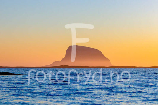 Lovund i solnedgang med hav og gyllent lys, majestetisk fjellprofil i horisonten.