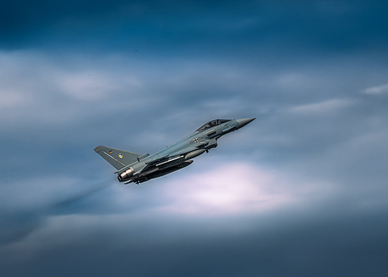 Eurofighter Typhoon i stigning mot dramatisk blågrå himmel, symboliserer fart og kraft.