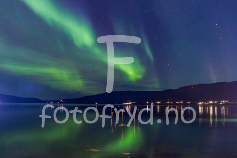 Nordlys som danser over stille fjord under stjerneklar himmel, med refleksjoner i vannet.
